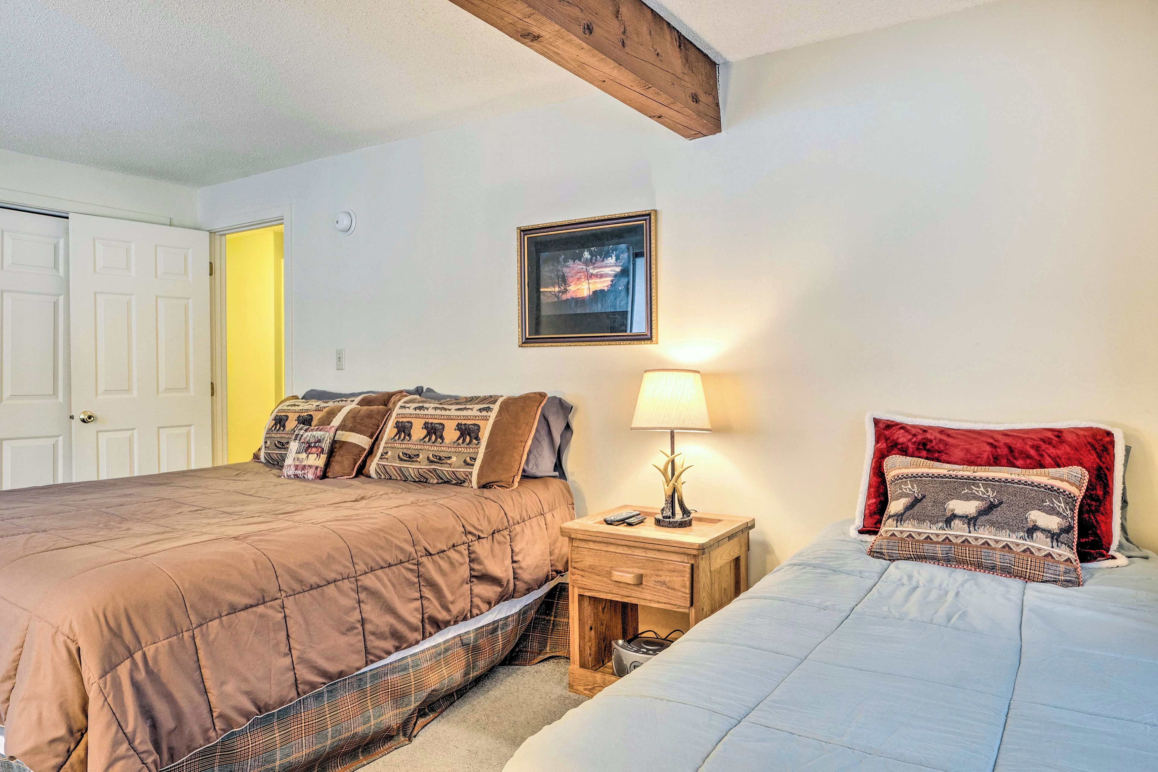 A bedroom with two beds, a nightstand with a lamp, a framed picture on the wall, and a door leading to another room.