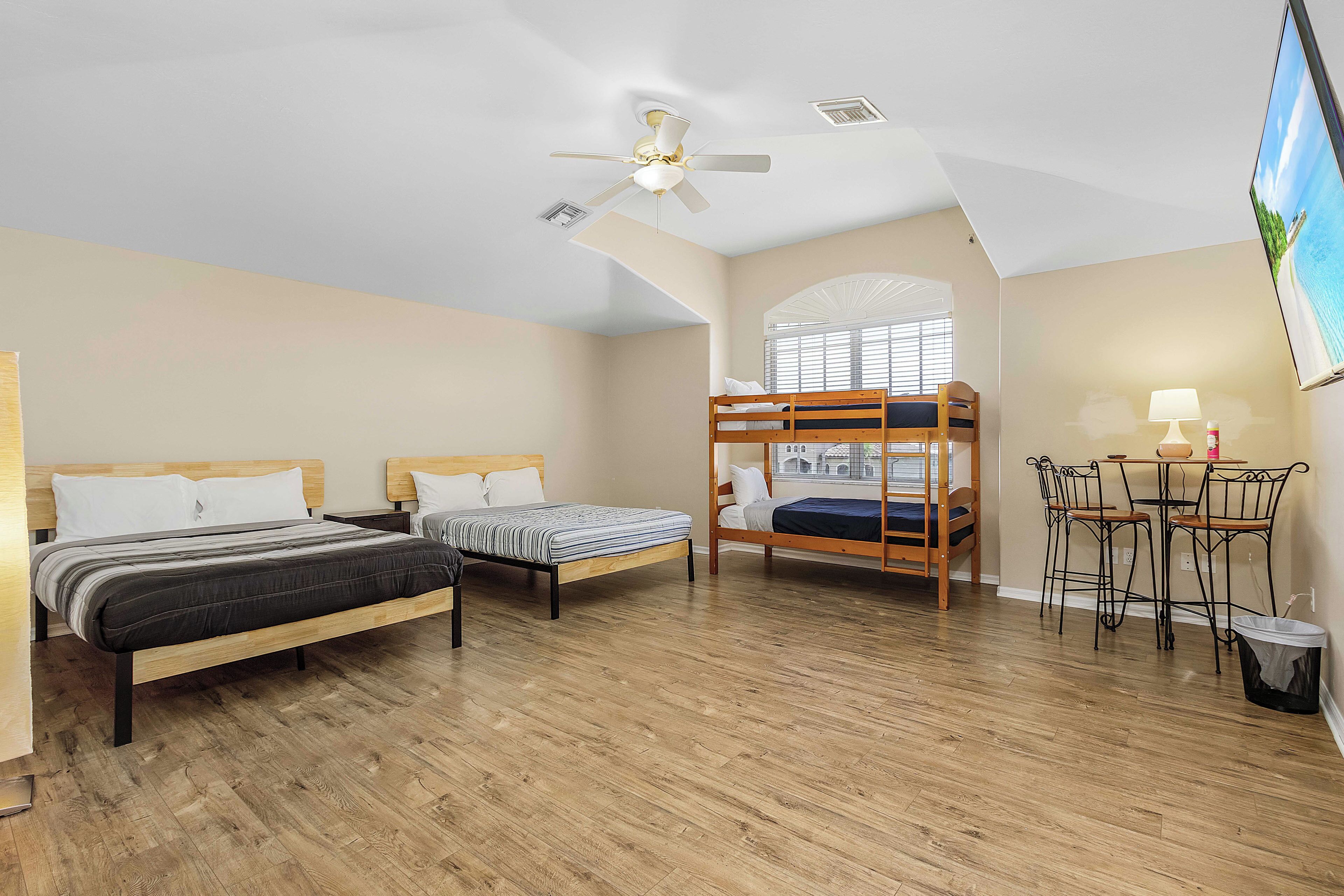 A bedroom with two beds, a ceiling fan, a window with blinds, a table with chairs, and a TV mounted on the wall.