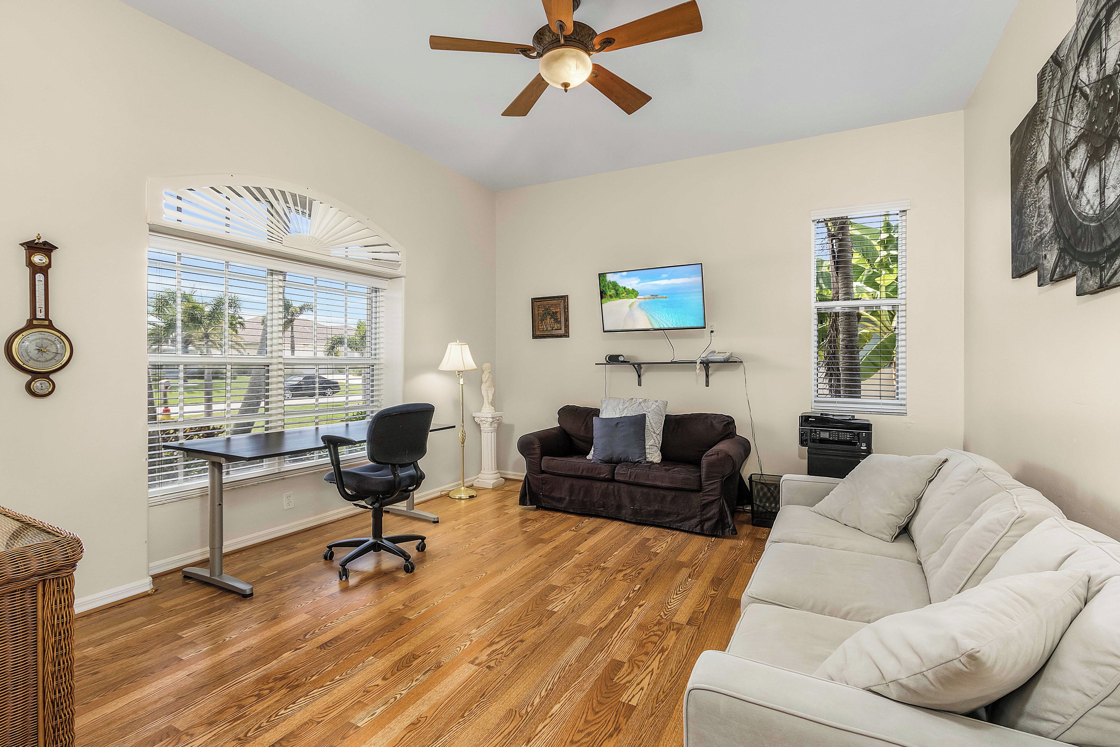 A living room with a wooden floor, a ceiling fan, a TV, a couch, a desk with a chair, and large windows with blinds.