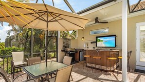 An outdoor patio with a glass table, chairs, a barbecue grill, and a TV screen displaying a beach scene.