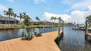 A wooden dock with two green Adirondack chairs, a glass-enclosed building, and a marina with boats.