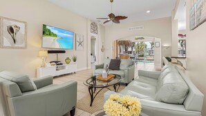 A spacious living room with a large flat-screen TV, a ceiling fan, and a sign reading "BEACH HOUSE".