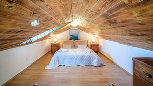 Une chambre confortable située sous les toits, avec un plafond en bois, un lit recouvert d’une courtepointe blanche, deux tables de chevet avec des lampes et un tableau accroché au mur.