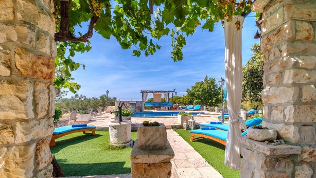 Une arche en pierre encadre un espace piscine agrémenté de chaises longues et d’une pergola.