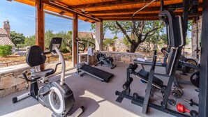 Une salle de sport en plein air équipée d’appareils d’exercice, notamment un vélo elliptique, un tapis de course et des appareils de musculation, située sous une pergola en bois offrant une vue sur les arbres et les bâtiments.
