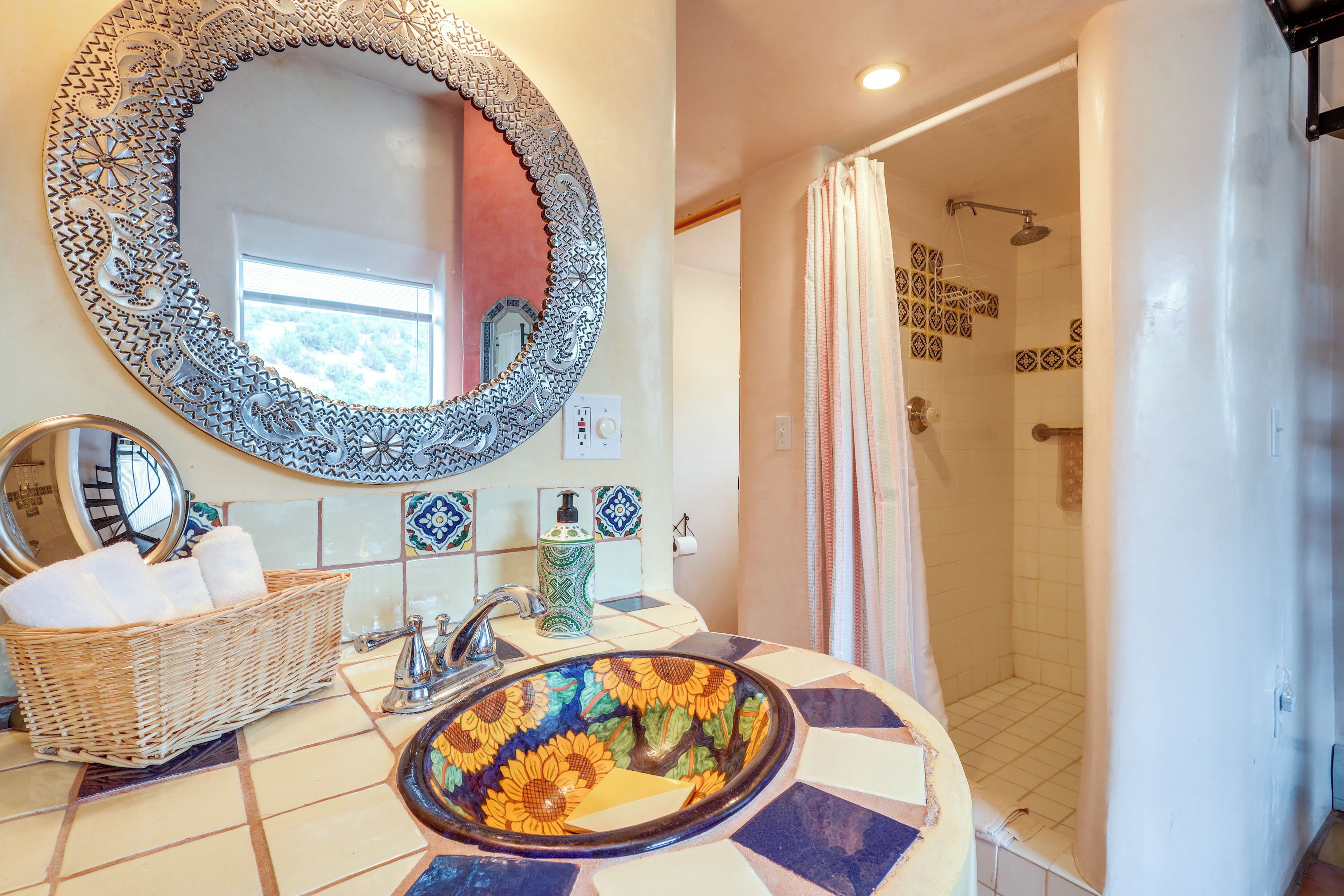 Une salle de bain avec un miroir décoratif, un lavabo surmonté d’une cuvette ornée de motifs de tournesols, un panier en osier rempli de serviettes et une douche aux carreaux décorés.