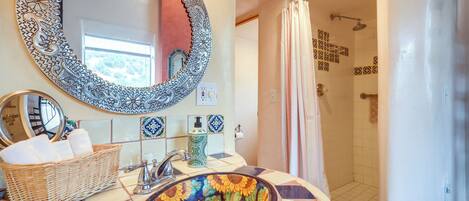 Une salle de bain avec un miroir décoratif, un lavabo surmonté d’une cuvette ornée de motifs de tournesols, un panier en osier rempli de serviettes et une douche aux carreaux décorés.
