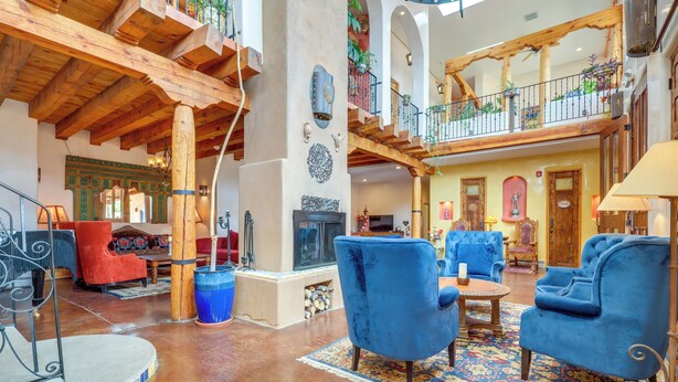 A spacious lobby with high ceilings, wooden beams, and a central fireplace. There are multiple seating areas with blue upholstered chairs and a red armchair. A large, ornate mirror hangs on the wall, and a staircase with a balcony is visible in the background. - Los Cerrillos Hillside Hacienda on 65 Acres! (Cerrillos)