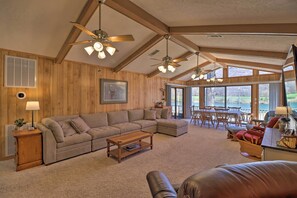 A spacious living room with a sectional sofa, a coffee table, and a leather couch. There are ceiling fans, a dining area with chairs, and large windows offering a view of the outdoors. - Scenic Riverview Getaway w/ Screened Porch! (Norfork)