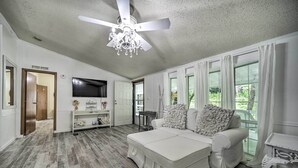 A spacious living room with a white sofa, a ceiling fan, and a large window with curtains. - Tranquil Creekfront Bryson City Home w/ Grill! (Bryson City)