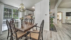 A spacious dining area with a large wooden table and chairs, a chandelier, and a view of the outdoors through large windows. - Tranquil Creekfront Bryson City Home w/ Grill! (Bryson City)