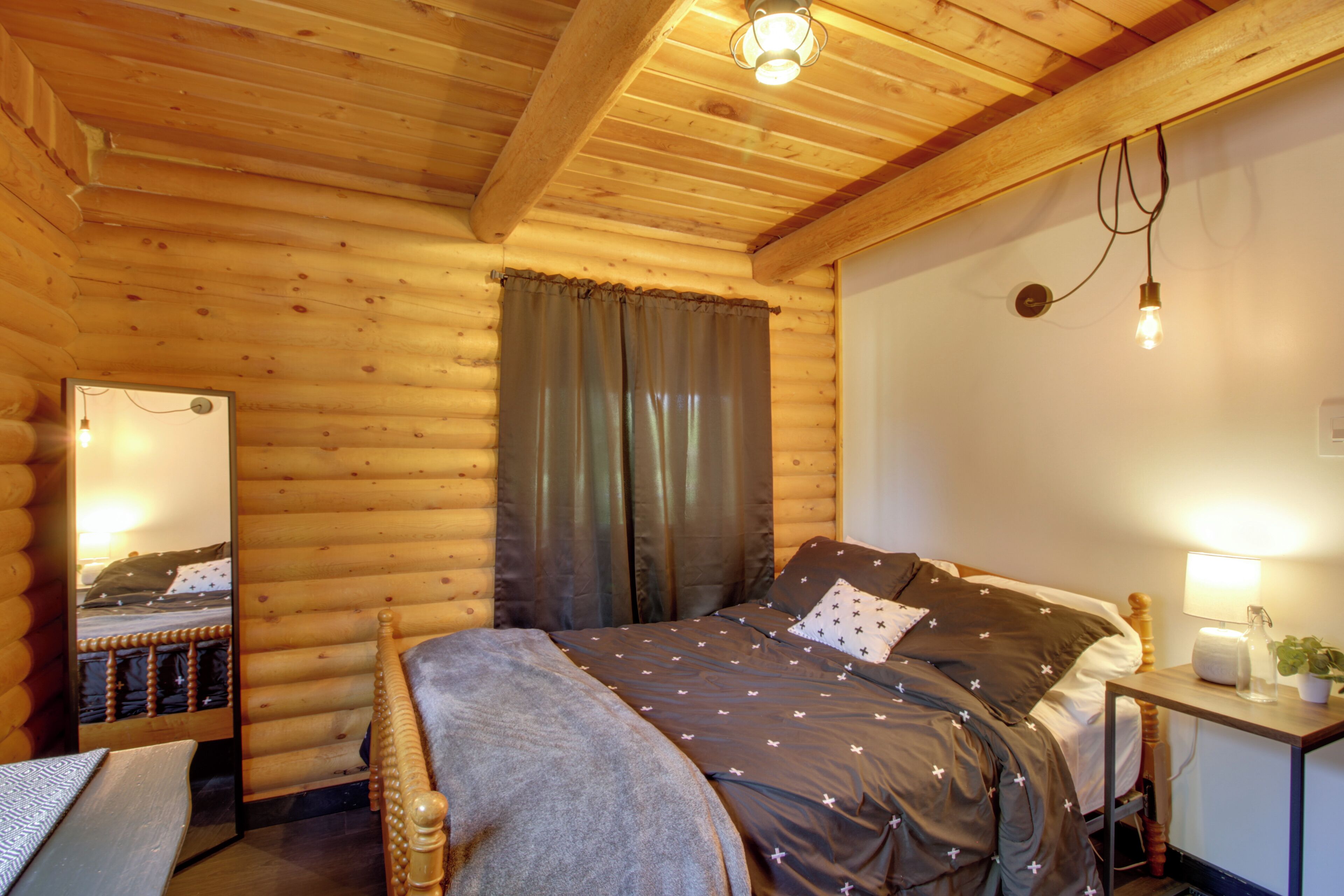 A cozy log cabin bedroom with a wooden bed, a bedside table with a lamp, and a mirror reflecting the room.