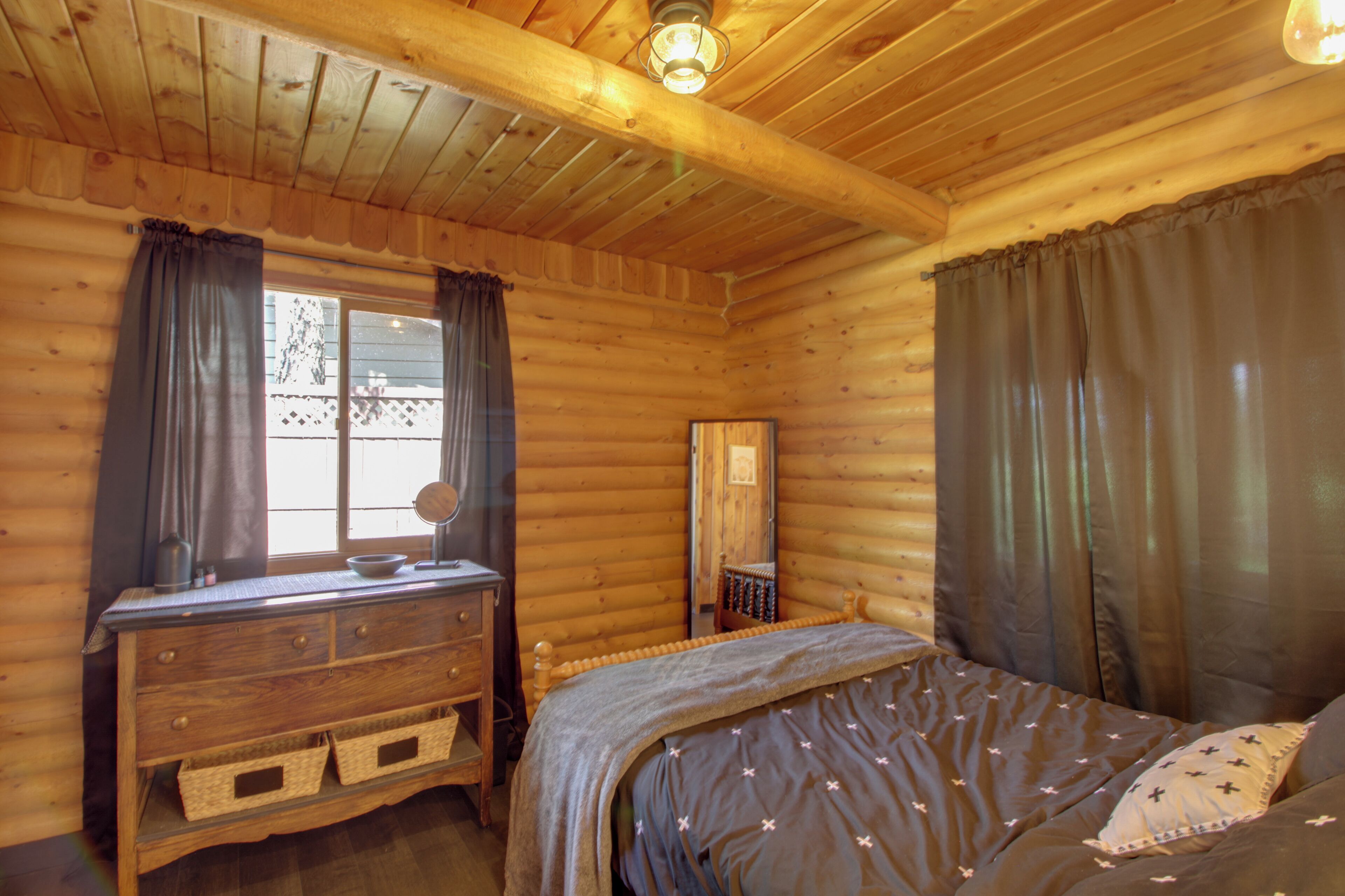 A wooden cabin bedroom with a bed, dresser, and window with curtains.