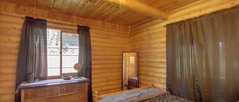 A wooden cabin bedroom with a bed, dresser, and window with curtains.