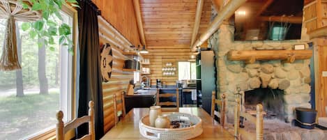 A wooden cabin interior with a stone fireplace, wooden dining table, and a hanging plant.