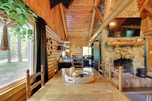 Interior de una cabaña de madera con chimenea de piedra, mesa de comedor de madera y una planta colgante.