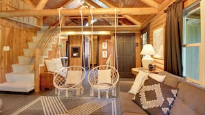 Ein rustikaler Innenraum einer Blockhütte mit einer Treppe, zwei Hängestühlen, einem Sofa und einer Lampe.