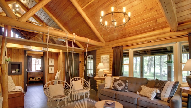 Ein Interieur einer Blockhütte mit Holzbalkendecke, einer Couch, zwei Korbstühlen, einem runden Couchtisch und einer Kandelaber.