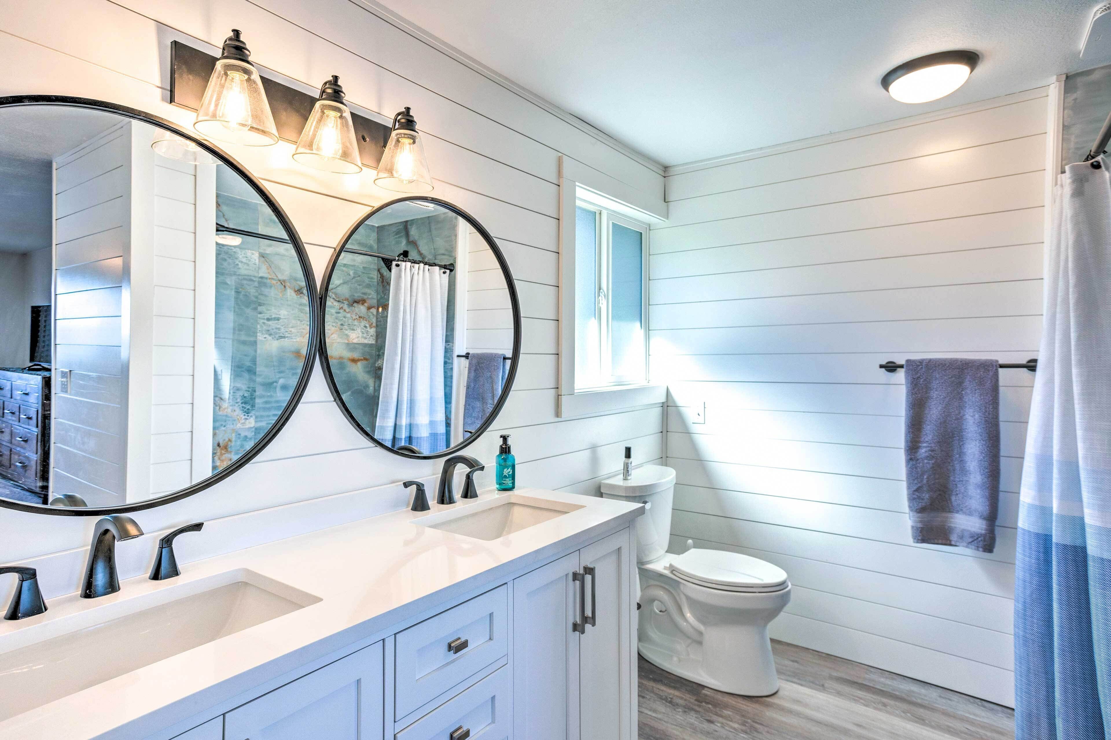 A bathroom with two sinks, a toilet, a mirror with lights, and a window.