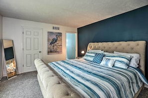 A bedroom with a large bed, a bench, a door, and a painting on the wall. - Charming Pentwater Home w/ Fire Pit & Yard! (Pentwater)