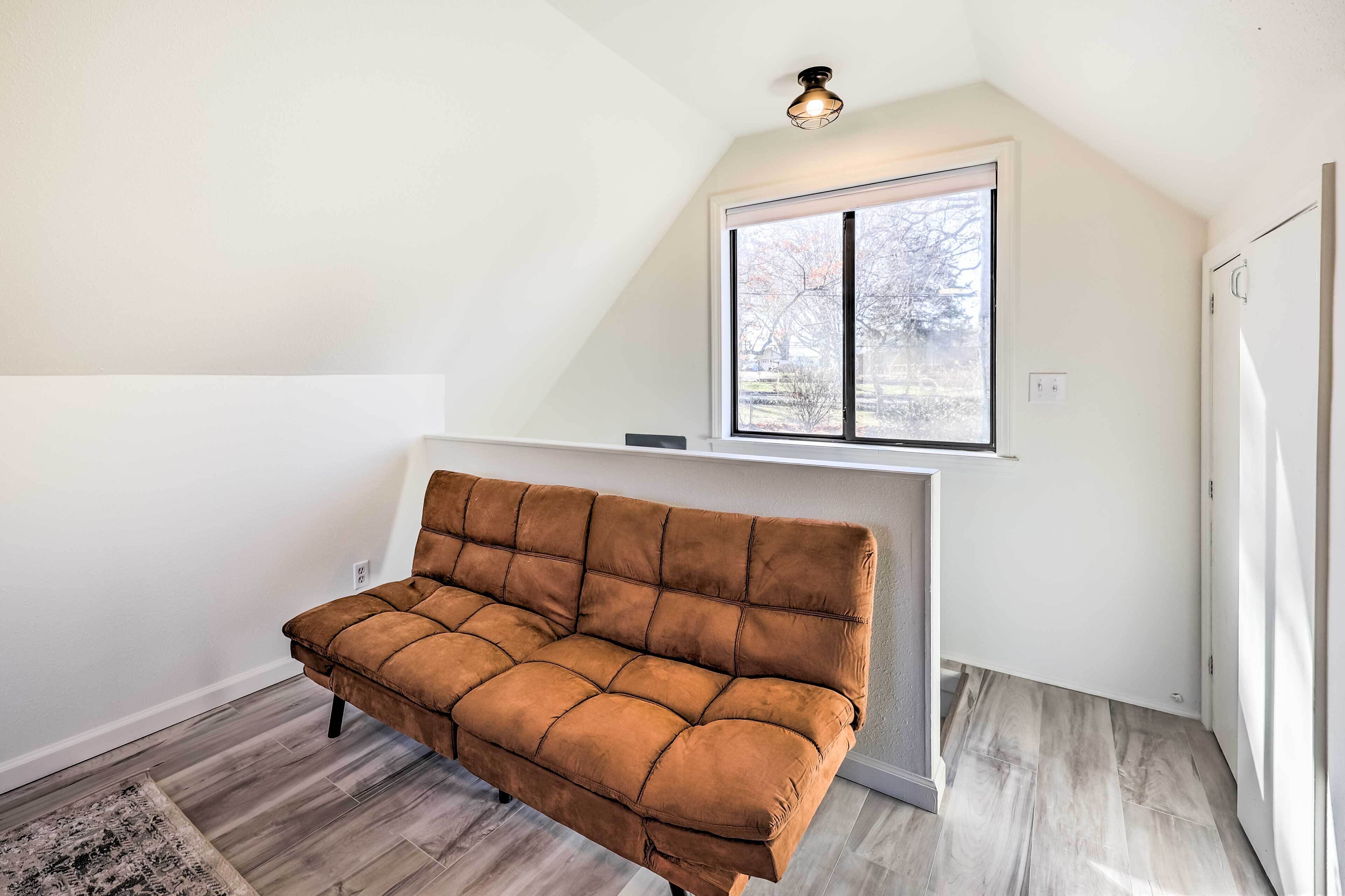 A room with a brown sofa, a white fireplace, and a large window with a view of trees.