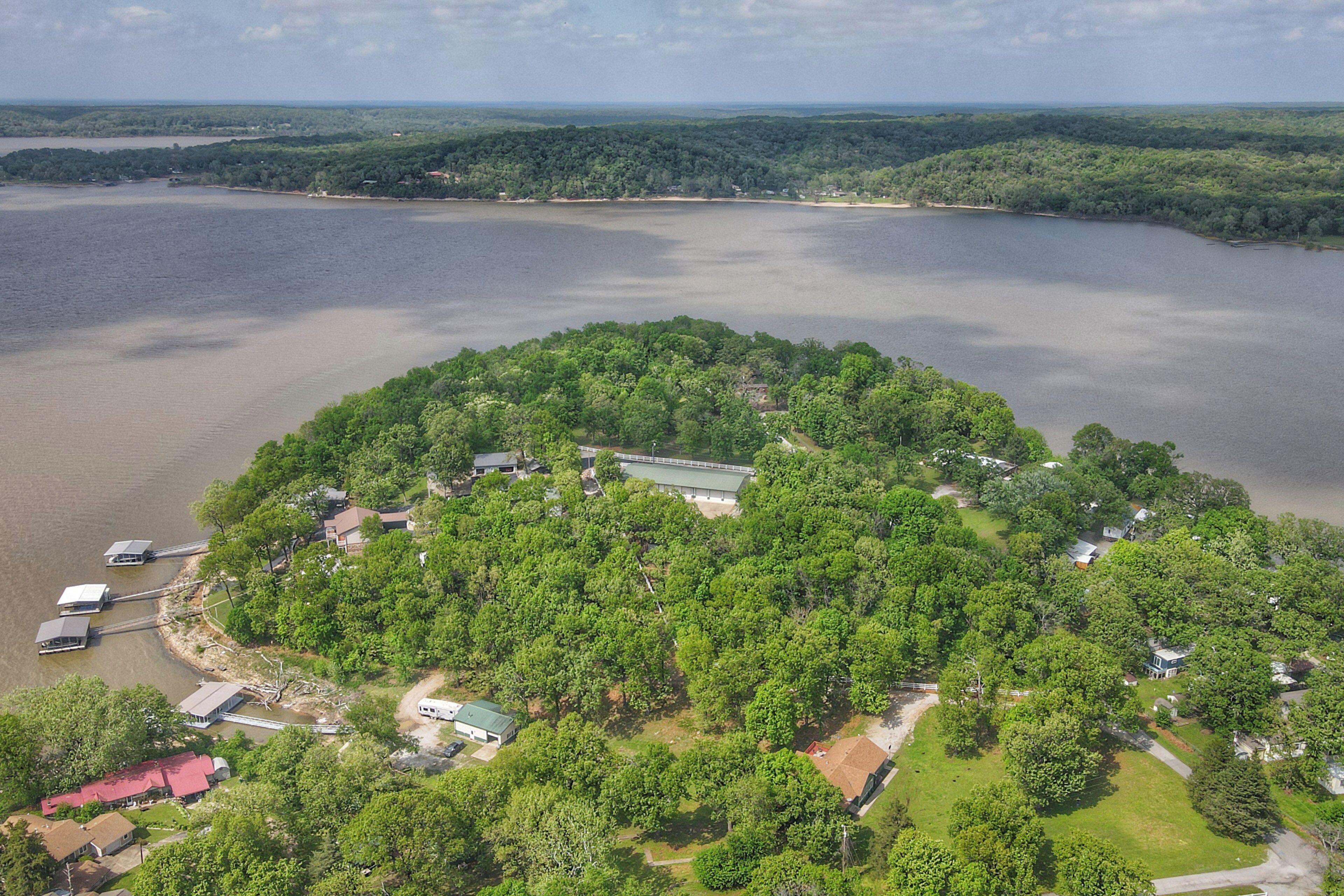 An aerial view of a lakeside property with a dock, several buildings, and dense greenery.