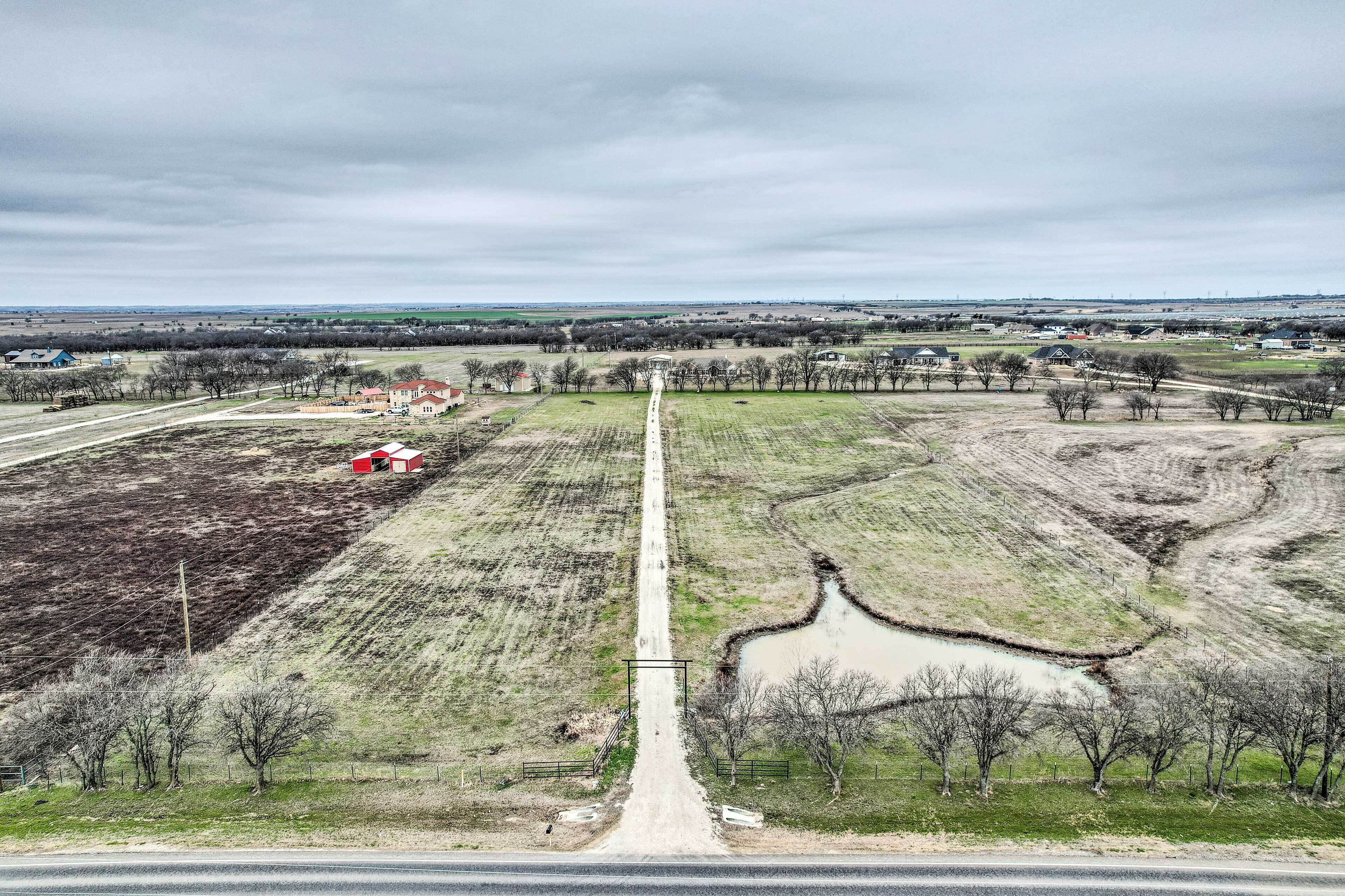Vista aérea de un paisaje rural con una carretera recta que conduce a un edificio rojo, rodeado de campos y árboles dispersos.
