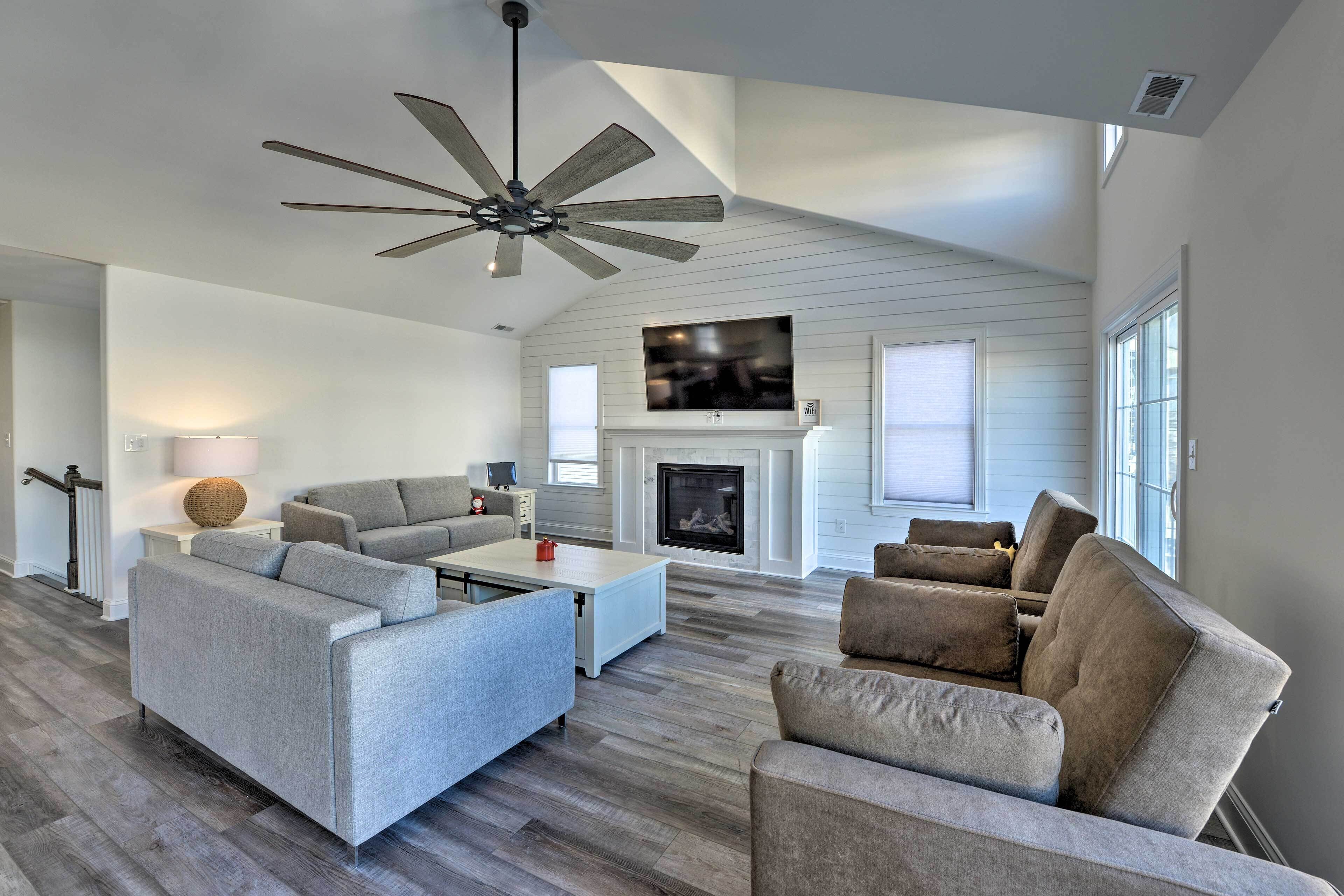 A spacious living room with a fireplace, a ceiling fan, and a large TV mounted above it.
