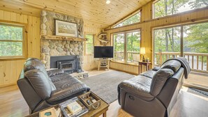 A cozy cabin interior with a stone fireplace, leather sofas, a flat-screen TV, and large windows offering a view of the forest. - Four-season Lake Cabin w/ Canoe & Paddle Boat! (Gordon)