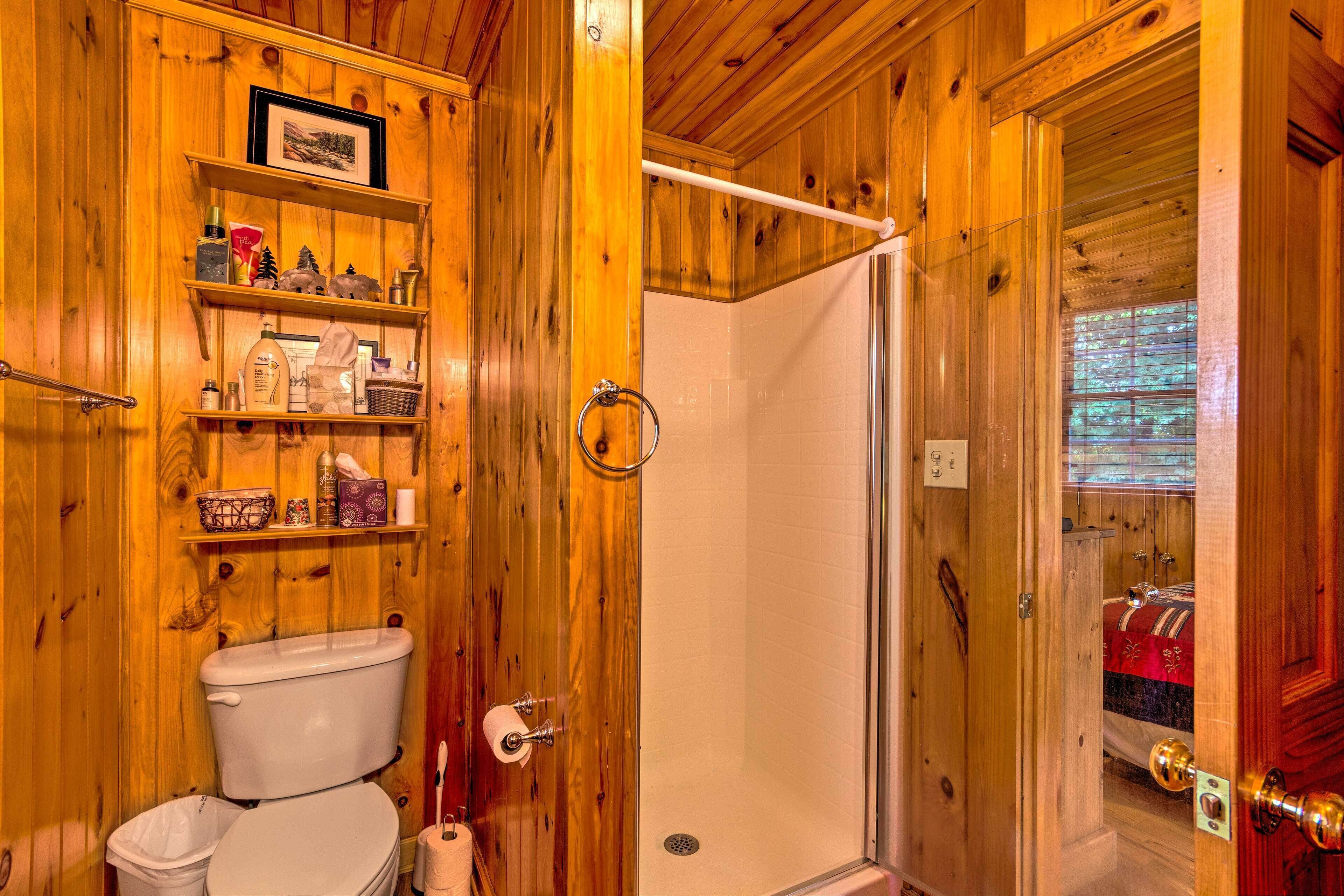 Interior de madeira com vaso sanitário, prateleiras com artigos de higiene, chuveiro e porta que dá para outro quarto.