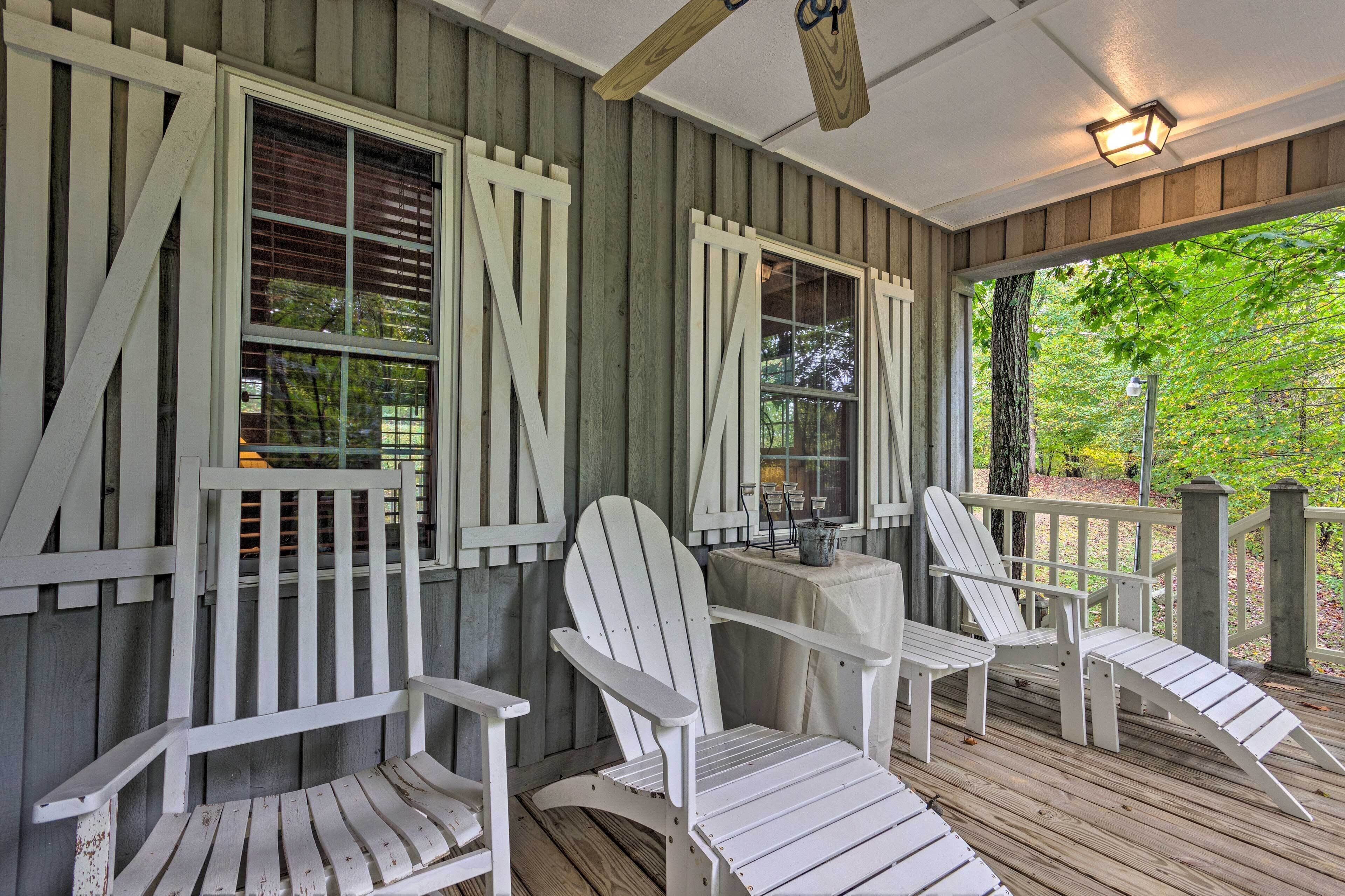 A covered porch with Adirondack chairs and a table, wooden walls, and a view of trees.
