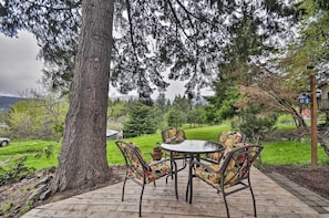 An outdoor patio with a round table and chairs, surrounded by a large tree and a view of a grassy area with a house in the background.
