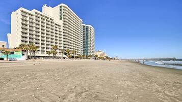 A beachfront hotel with multiple floors, a sandy beach in the foreground, and a pier extending into the ocean in the background.
