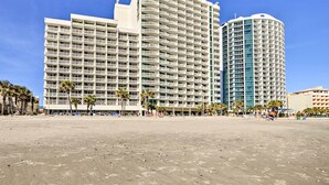 A beachfront hotel with multiple balconies, a sandy beach in the foreground, and palm trees. - Oceanfront Myrtle Beach Condo w/ Balcony! (Myrtle Beach)