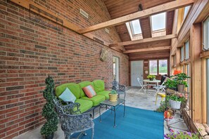 A covered patio with a brick wall, wooden beams, a green sofa, metal chairs, a small table, and potted plants. - Peaceful Fayetteville Getaway - Pets Welcome! (Fayetteville)