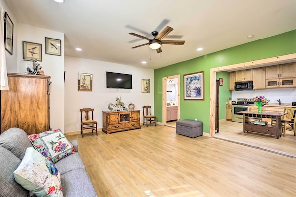 A living room with a sofa, a wooden cabinet, a ceiling fan, and a kitchen area with wooden cabinets and a microwave.