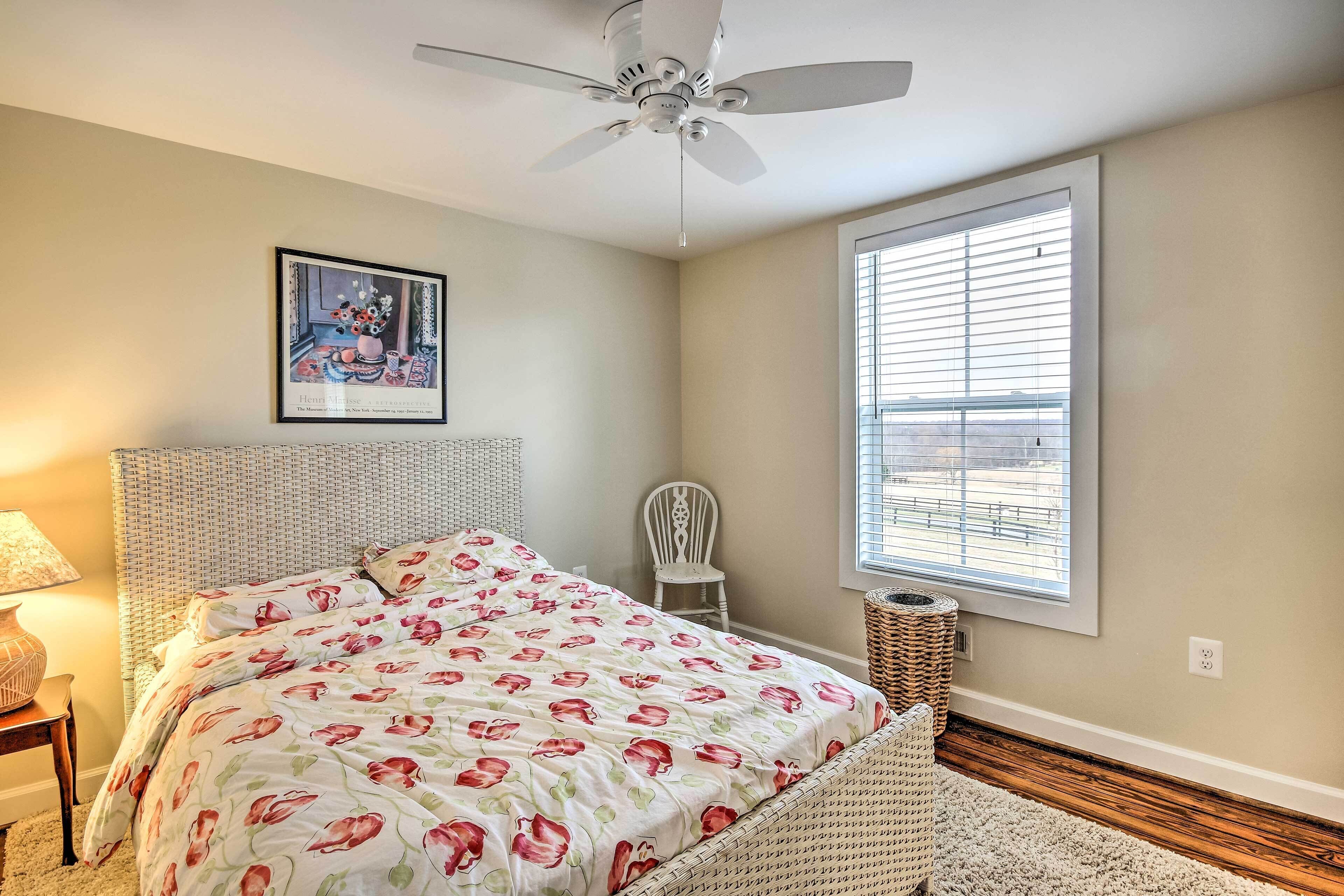 A bedroom with a bed, a ceiling fan, a window with blinds, a wicker chair, and a lamp.