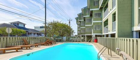 Una piscina con sillones de descanso, una valla y un edificio de varios pisos al fondo.