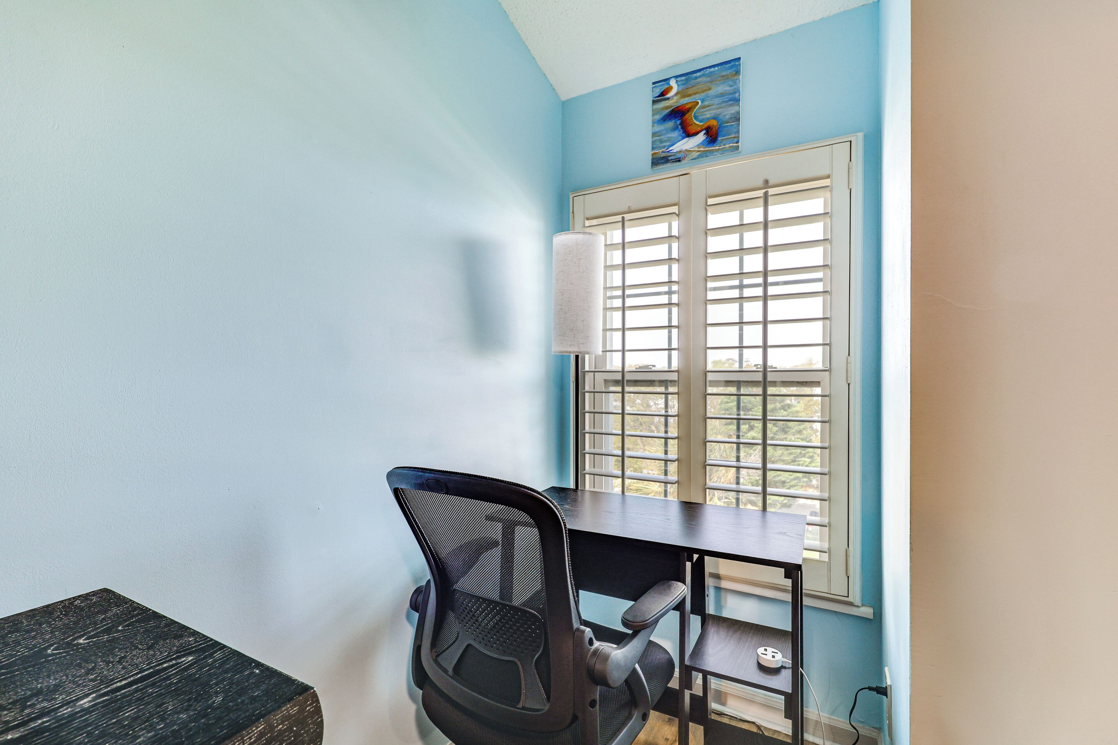 Una habitación con escritorio, silla, ventana con persianas y un cuadro en la pared.