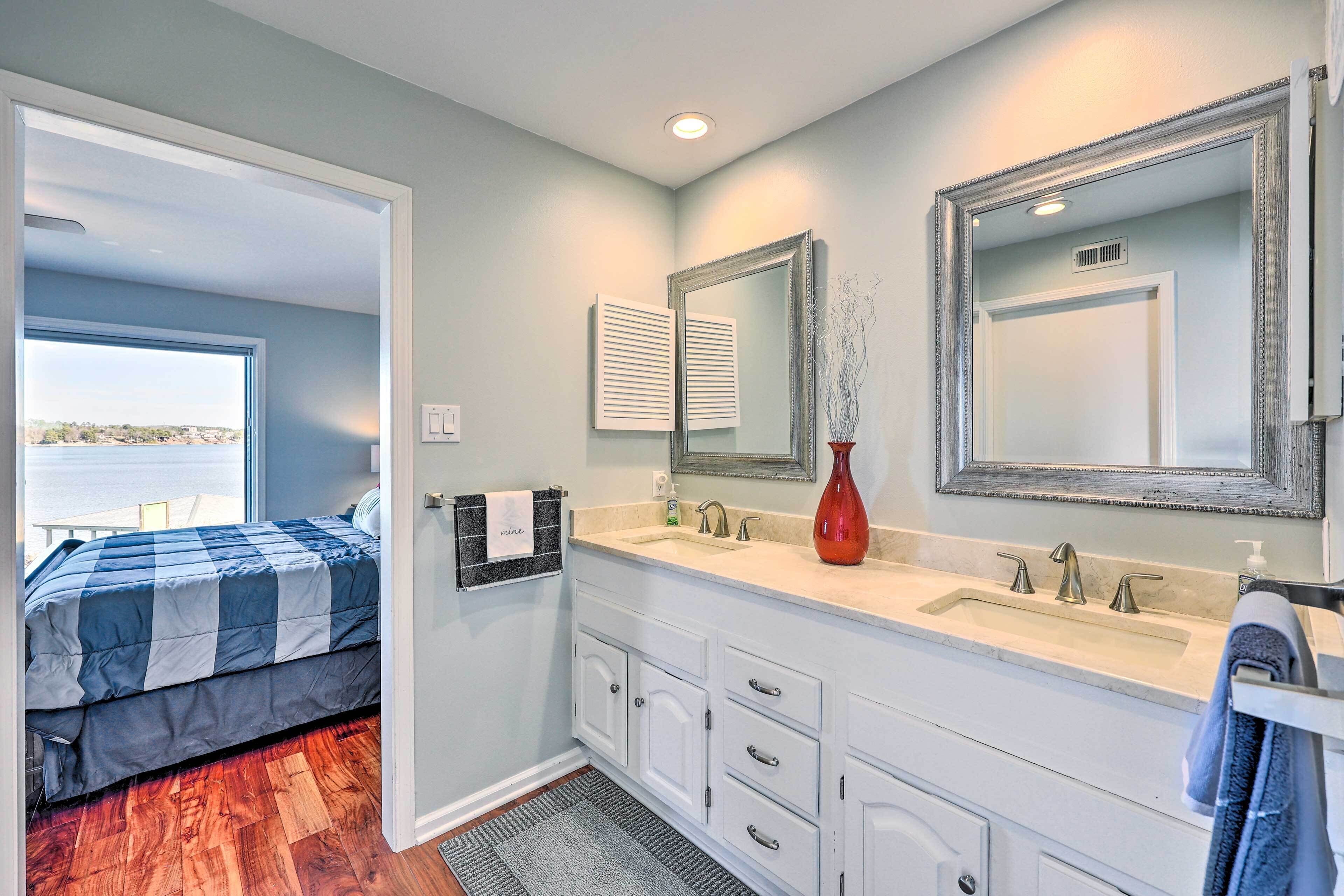 A bedroom with a large window, a bed with a blue and white striped blanket, a bathroom with a double sink, a mirror, and a red vase.