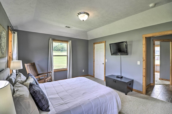 A bedroom with a large bed, a chair, a desk, a television, and a door leading to a balcony. - Mountain Home Cottage w/ Deck on White River! (Gassville)