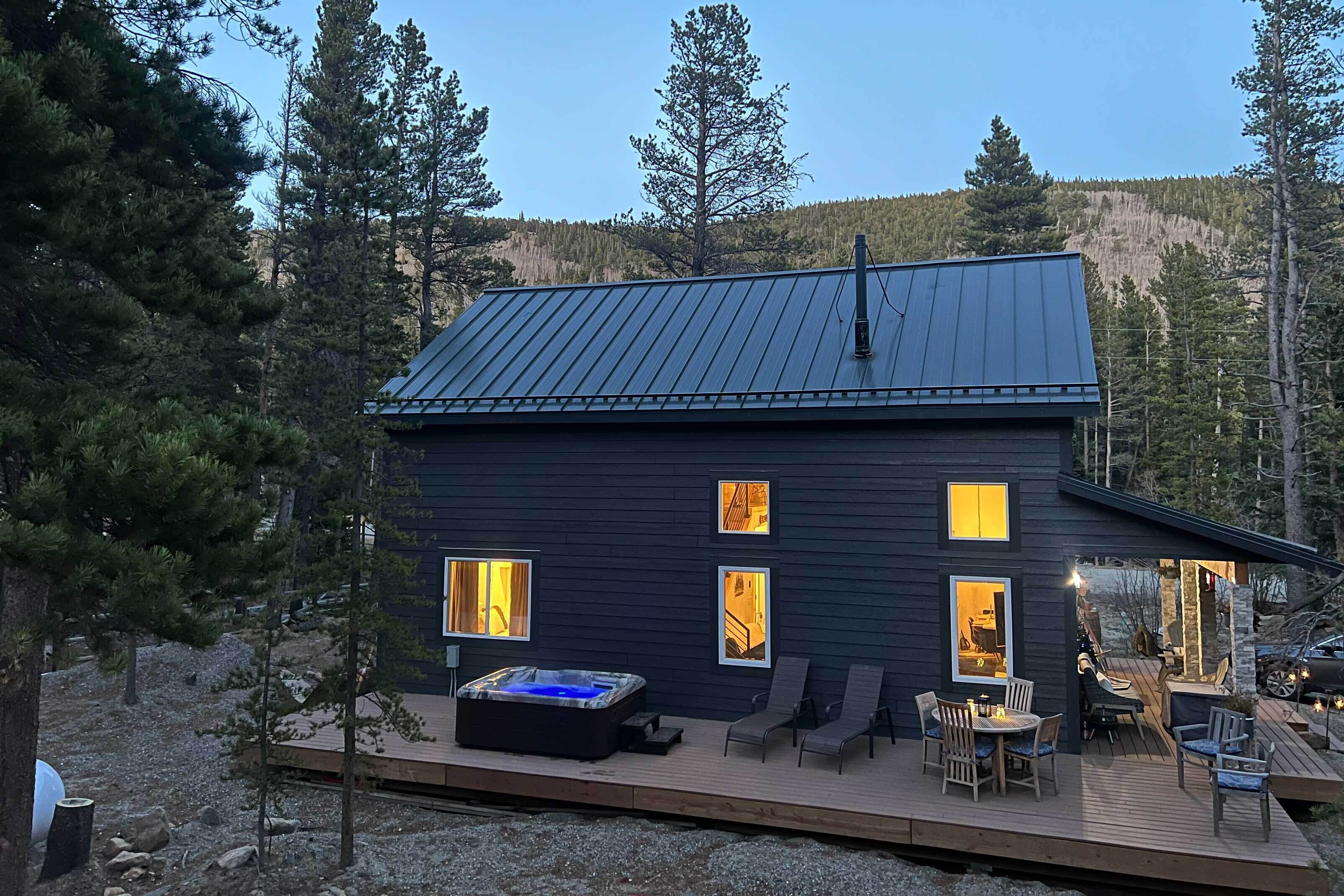 Una cabaña de madera oscura con techo metálico, un jacuzzi y una terraza con mobiliario exterior.