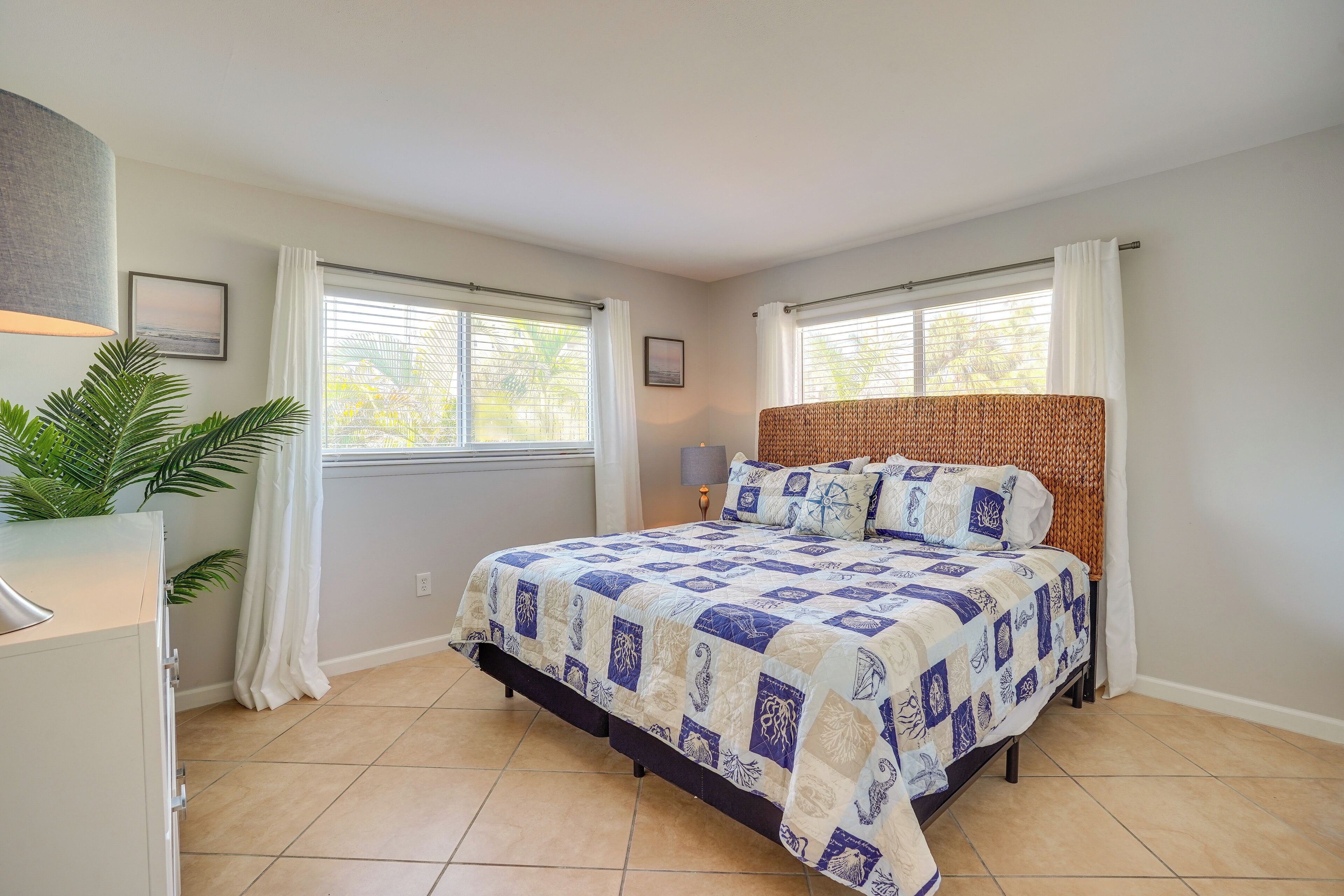 A bedroom with a bed, two windows, a nightstand, a lamp, and a potted plant.
