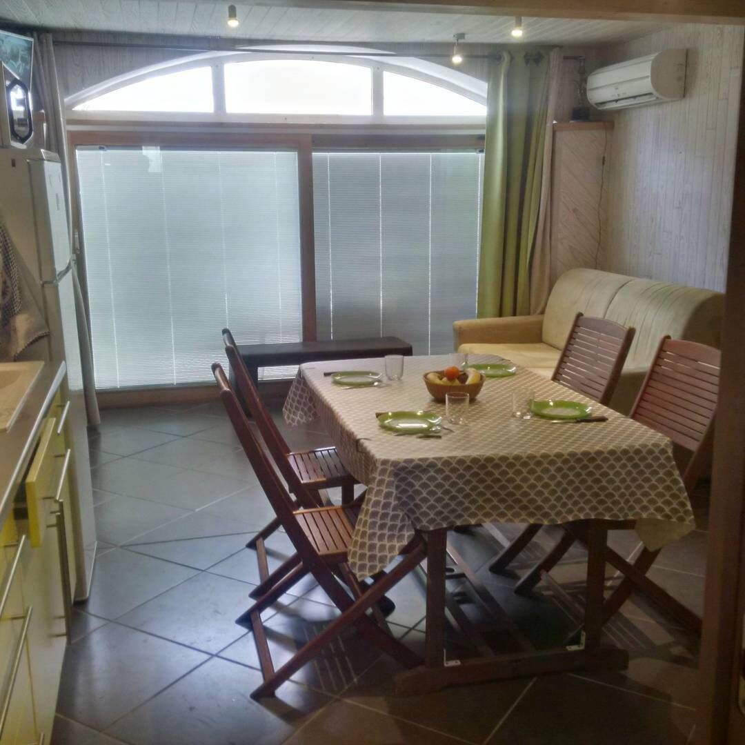 Un comedor con una mesa dispuesta para cuatro personas, rodeada de sillas de madera. Hay un sofá y una ventana con persianas.