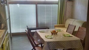 A dining area with a table set for four, surrounded by wooden chairs. There is a sofa and a window with blinds. - APT 2 ROOMS 27m2 + TERRACE 22m2 REF 4015 (MARSEILLAN PLAGE)