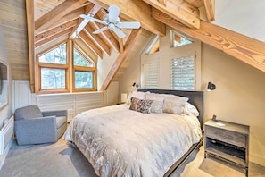 A bedroom with a sloped ceiling, a bed with a white duvet, a gray armchair, and a nightstand with a lamp. - Serene Sun Valley Home w/ Hot Tub & Mtn Views (Sun Valley)