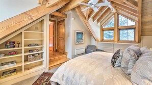 A bedroom with a sloped ceiling, wooden beams, a large bed, a window with a view of trees, and a bookshelf with games. - Serene Sun Valley Home w/ Hot Tub & Mtn Views (Sun Valley)