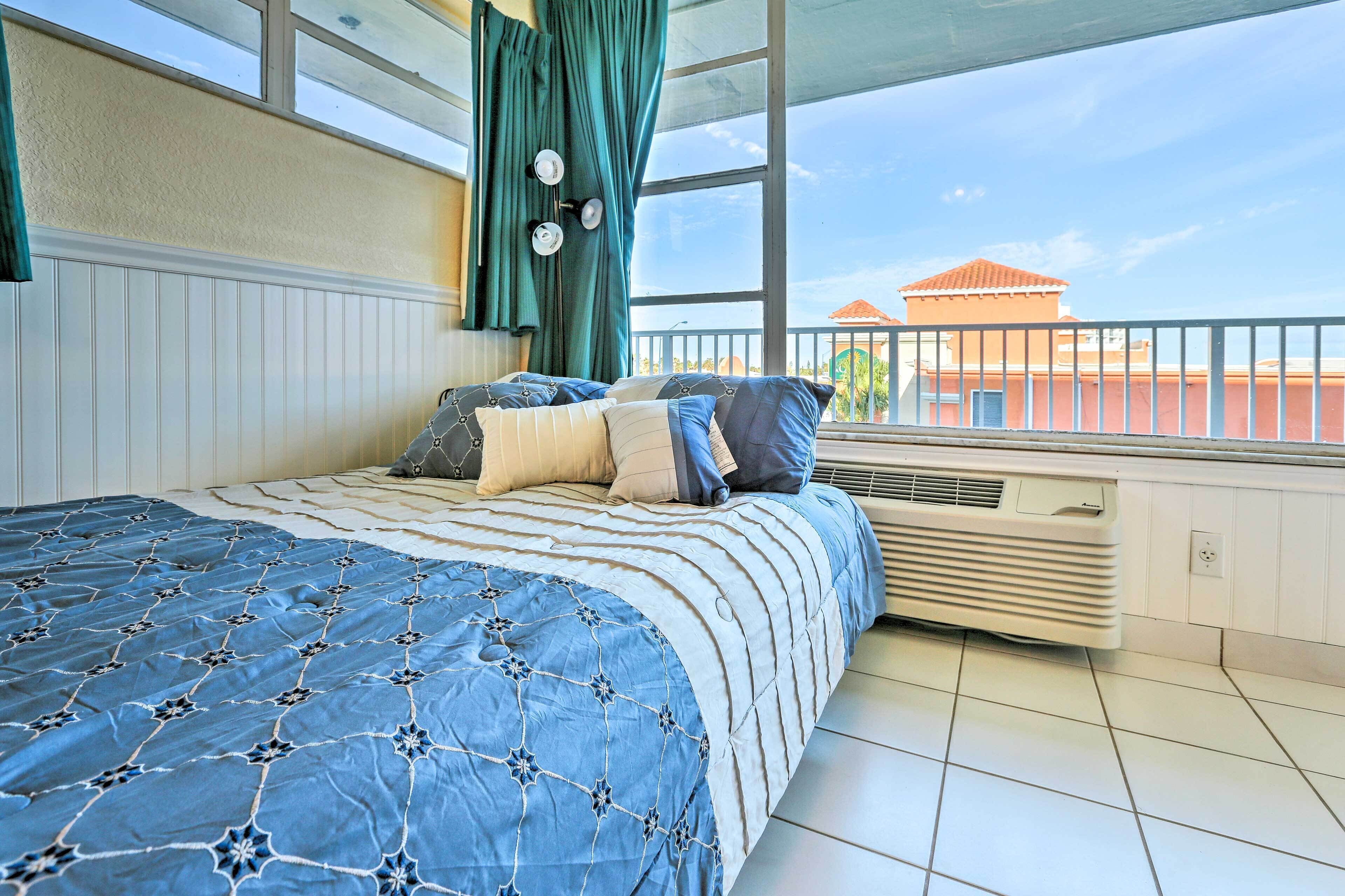 A bedroom with a large bed, a window with curtains, and a view of buildings outside.