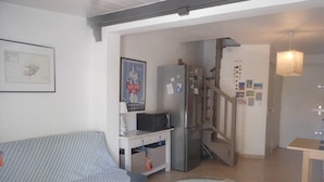 A living room with a sofa, a round glass table, a refrigerator, and a staircase leading to another level. - VILLA  3 ROOMS MEZ 47m2 + TERRACE 15m2 REF 4004 (MARSEILLAN PLAGE)