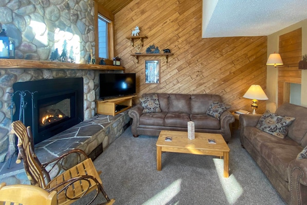 A cozy living room with a stone fireplace, a brown leather sofa, a wooden coffee table, and a television.